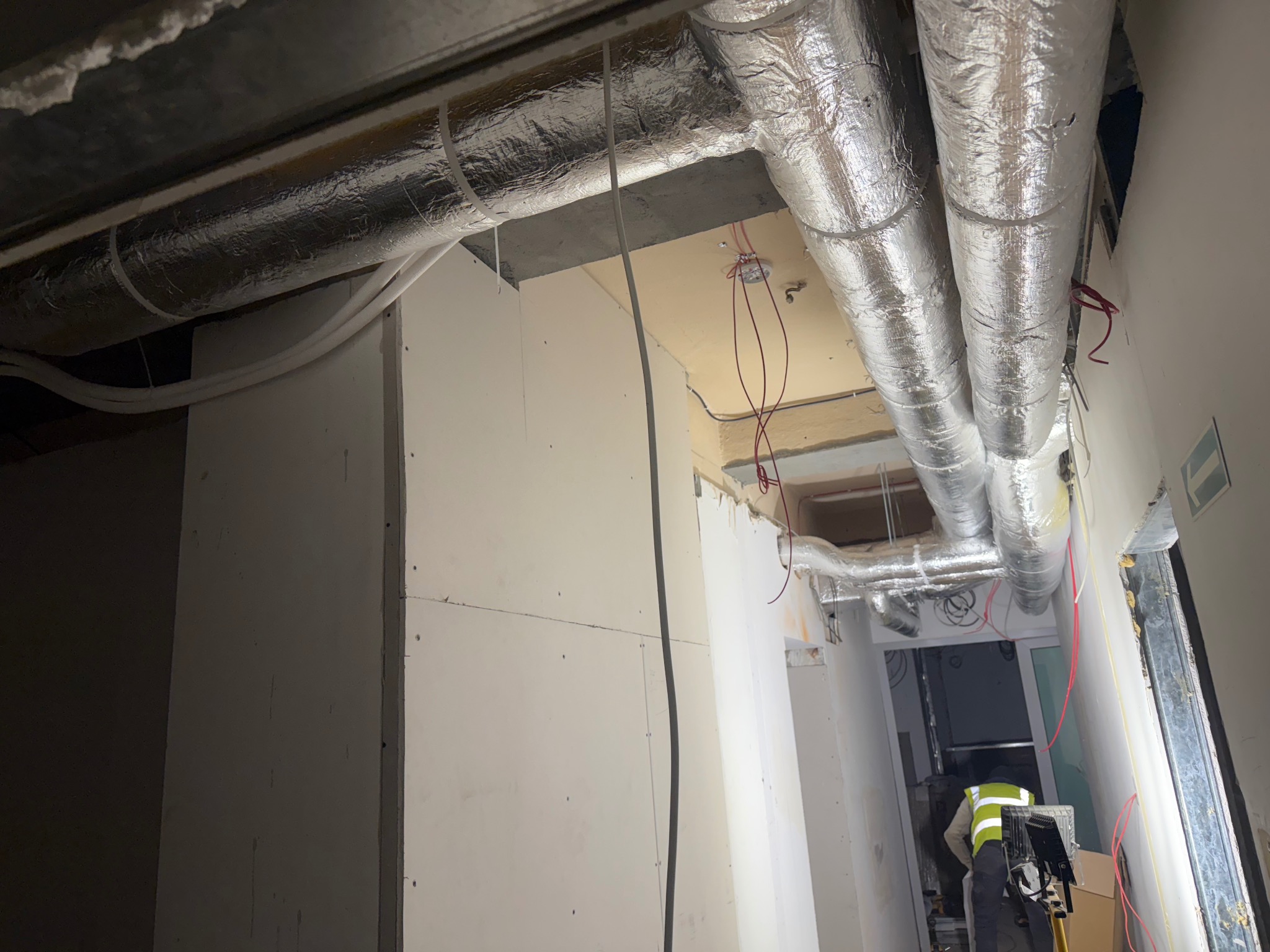 Montaż izolowanych przewodów wentylacyjnych pod sufitem, widoczne kable i fragment pracownika w kamizelce odblaskowej. Prace instalacyjne w budynku.