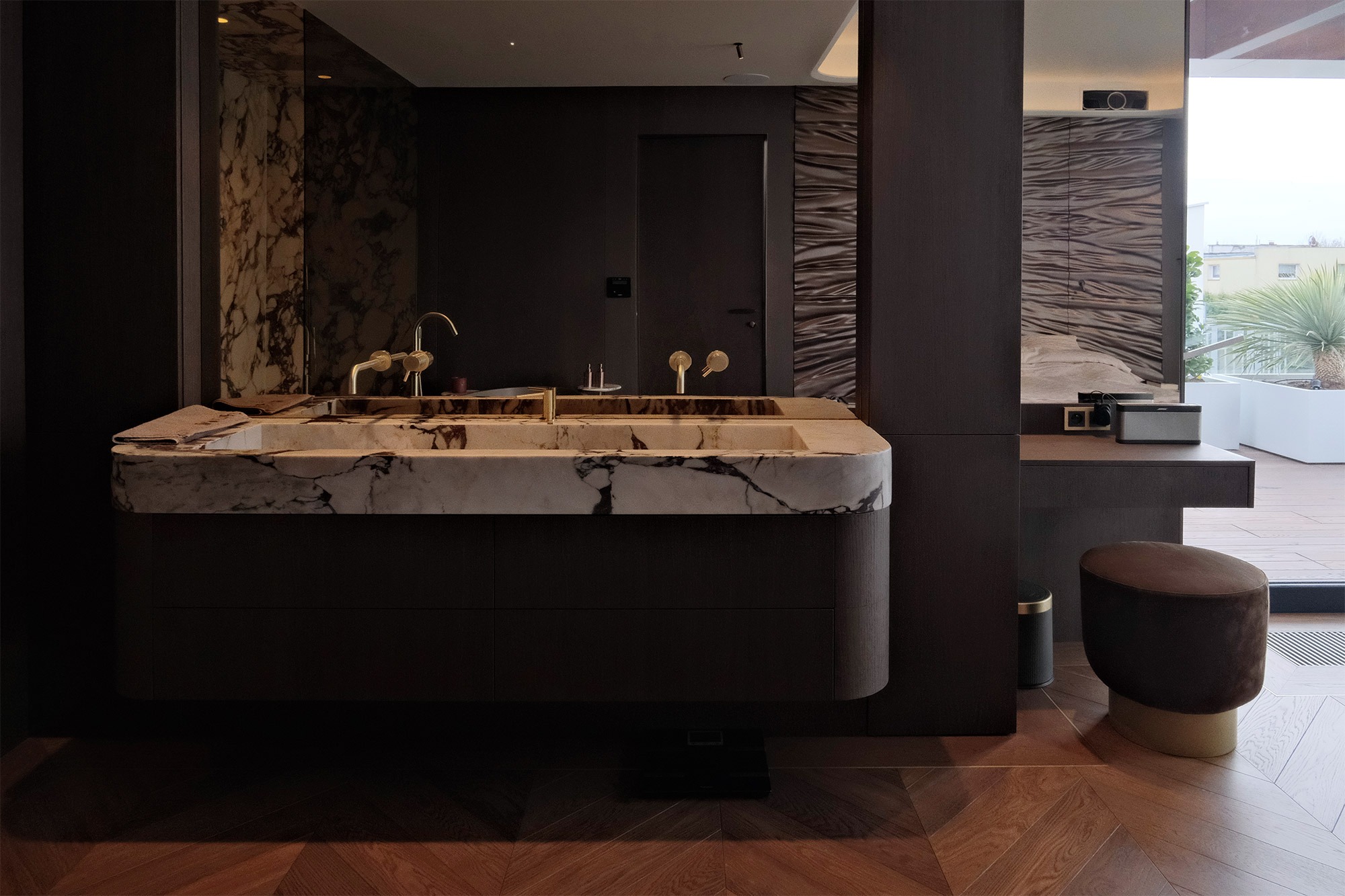 Nowoczesna łazienka z marmurową umywalką i złotą armaturą. Parkiet w jodełkę, ciemne meble i widok na taras z roślinami w tle. Minimalistyczny design.