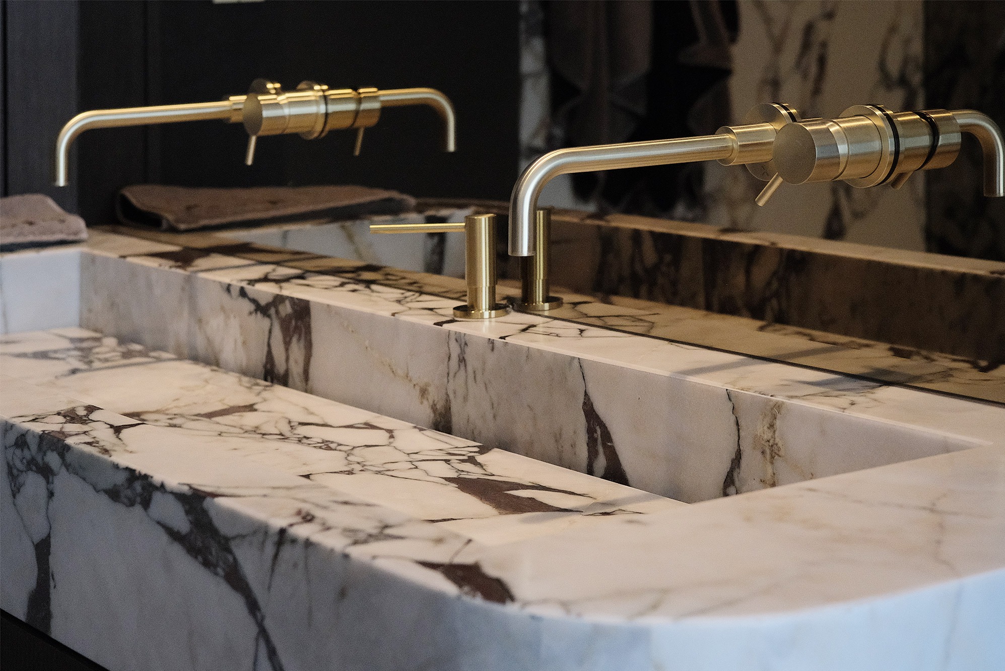 Marmurowa umywalka z dwoma złotymi bateriami o unikalnym designie. Widoczne detale marmuru i precyzyjne wykonanie armatury. Nowoczesny minimalizm w łazience.