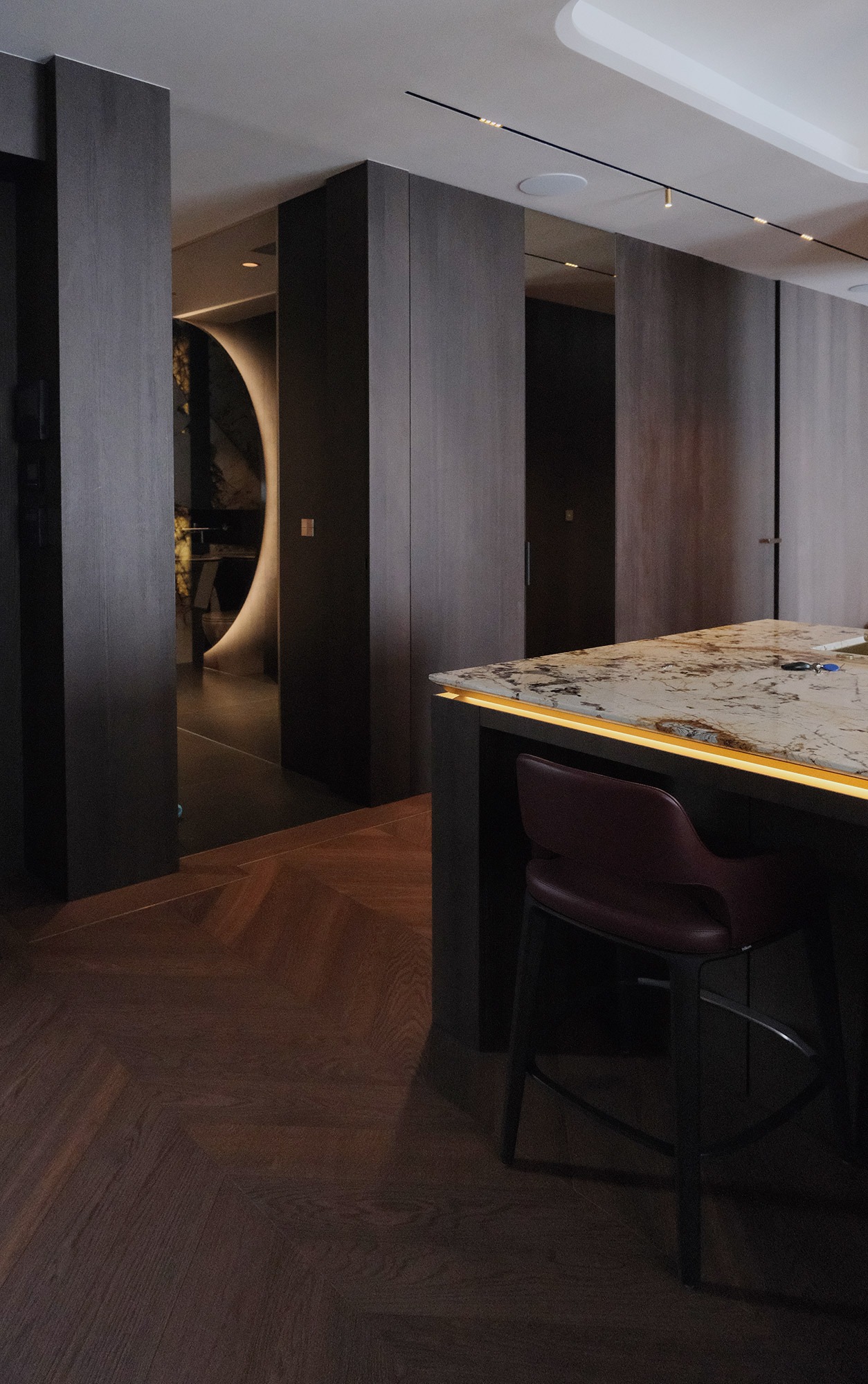 Nowoczesne wnętrze: parkiet w jodełkę, wyspa kuchenna z podświetleniem LED i krzesłem barowym. W tle widoczne drzwi z okrągłym oświetleniem.