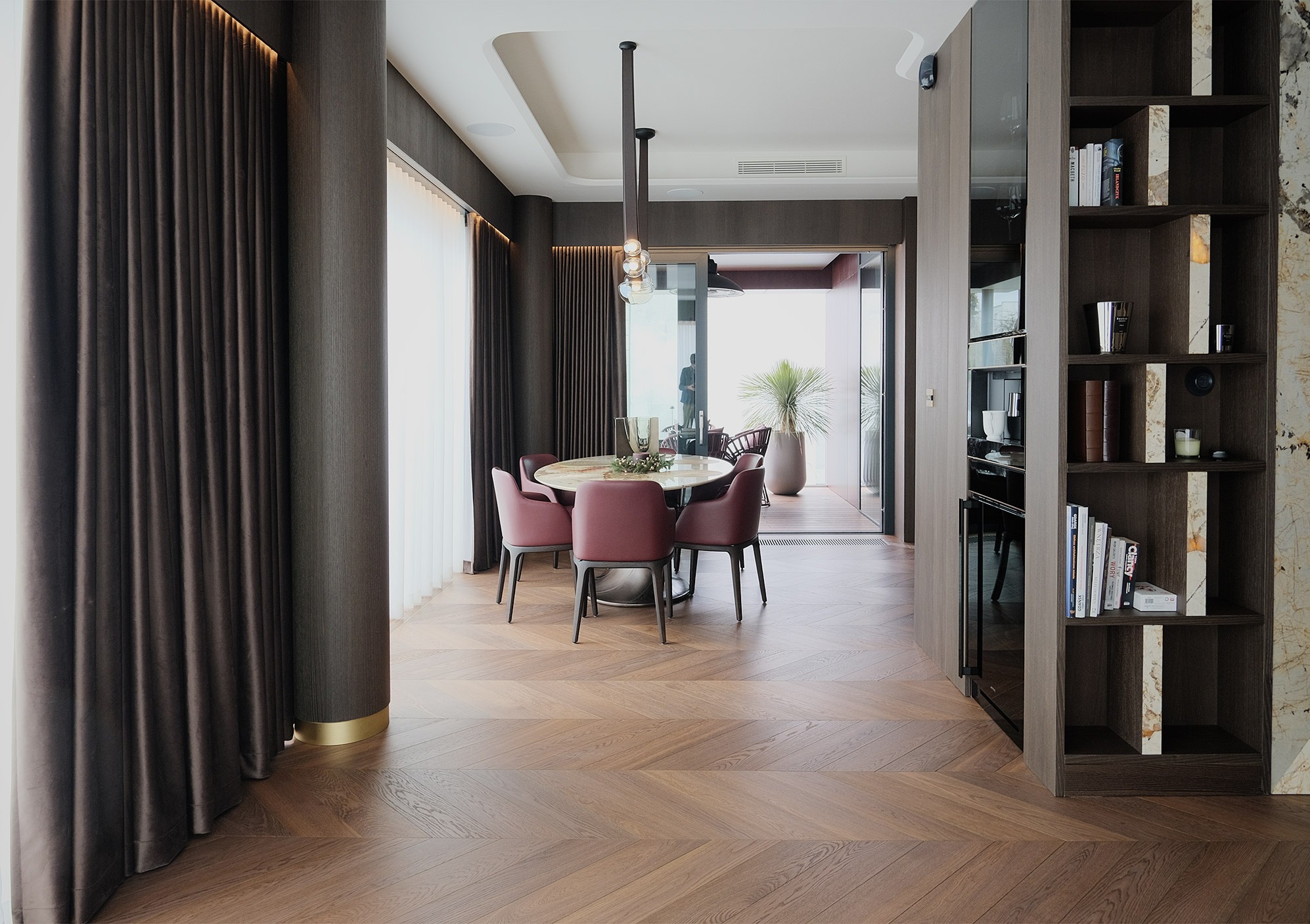 Elegancka jadalnia z parkietem w jodełkę, okrągłym stołem i krzesłami w kolorze burgundu. Widok na taras z roślinami. Nowoczesne oświetlenie i zabudowa meblowa.