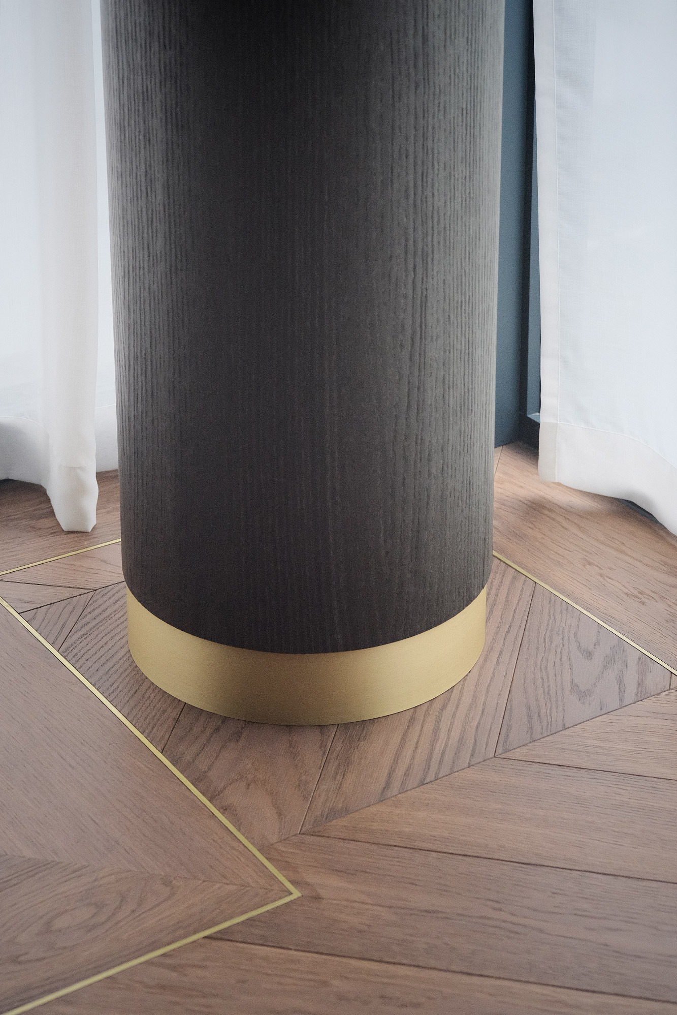 Parkiet w jodełkę z mosiężnymi intarsjami, otaczający ciemny, okrągły cokół z drewnianą fakturą i złotym wykończeniem, przy białej zasłonie.