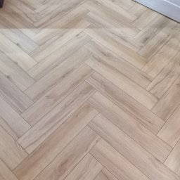 Dariusz Dymkowski - Jasne panele podłogowe ułożone w jodełkę, widoczne przy białej listwie przypodłogowej. Delikatny wzór drewna, ciepłe oświetlenie.