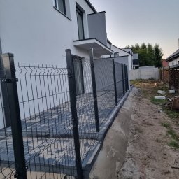 Usługi brukarskie OLBRUK OLEKSANDR STETS - Nowoczesne ogrodzenie panelowe w kolorze grafitowym, zamontowane przy posesji z kostką brukową. Widok na dom z jasną elewacją i fragment ogrodu.