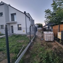 Usługi brukarskie OLBRUK OLEKSANDR STETS - Nowoczesne ogrodzenie panelowe z podmurówką na tle domu jednorodzinnego w trakcie montażu. Widoczny zbiornik na wodę i trawa.