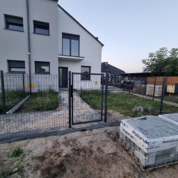 Usługi brukarskie OLBRUK OLEKSANDR STETS - Nowoczesny dom z szarą elewacją i metalowym ogrodzeniem panelowym. Częściowo ułożona kostka brukowa i paleta kostki Polbruk. Widok na ogród i zabudowania gospodarcze.