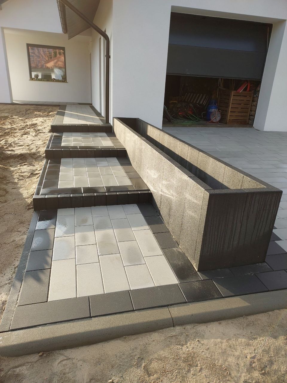 Nowoczesne schody wejściowe z kostki brukowej w odcieniach szarości i bieli, prowadzące do domu z garażem. Długa, prostokątna donica z boku schodów.