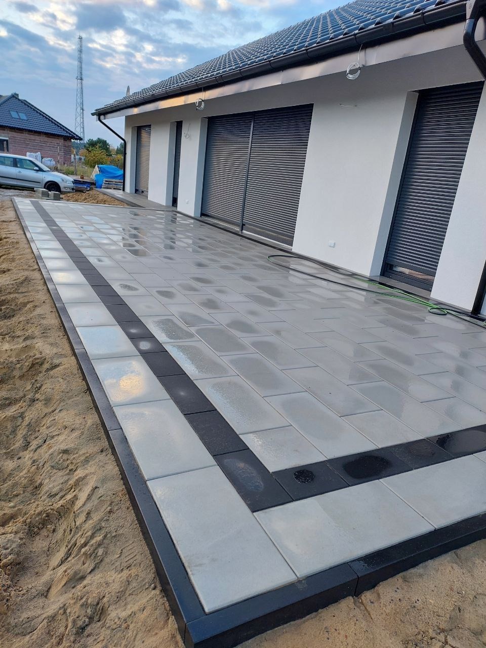 Ułożona, jasnoszara kostka brukowa z czarnymi obramowaniami na podjeździe domu z szarymi roletami. Widoczny piasek po bokach i fragment nieba.