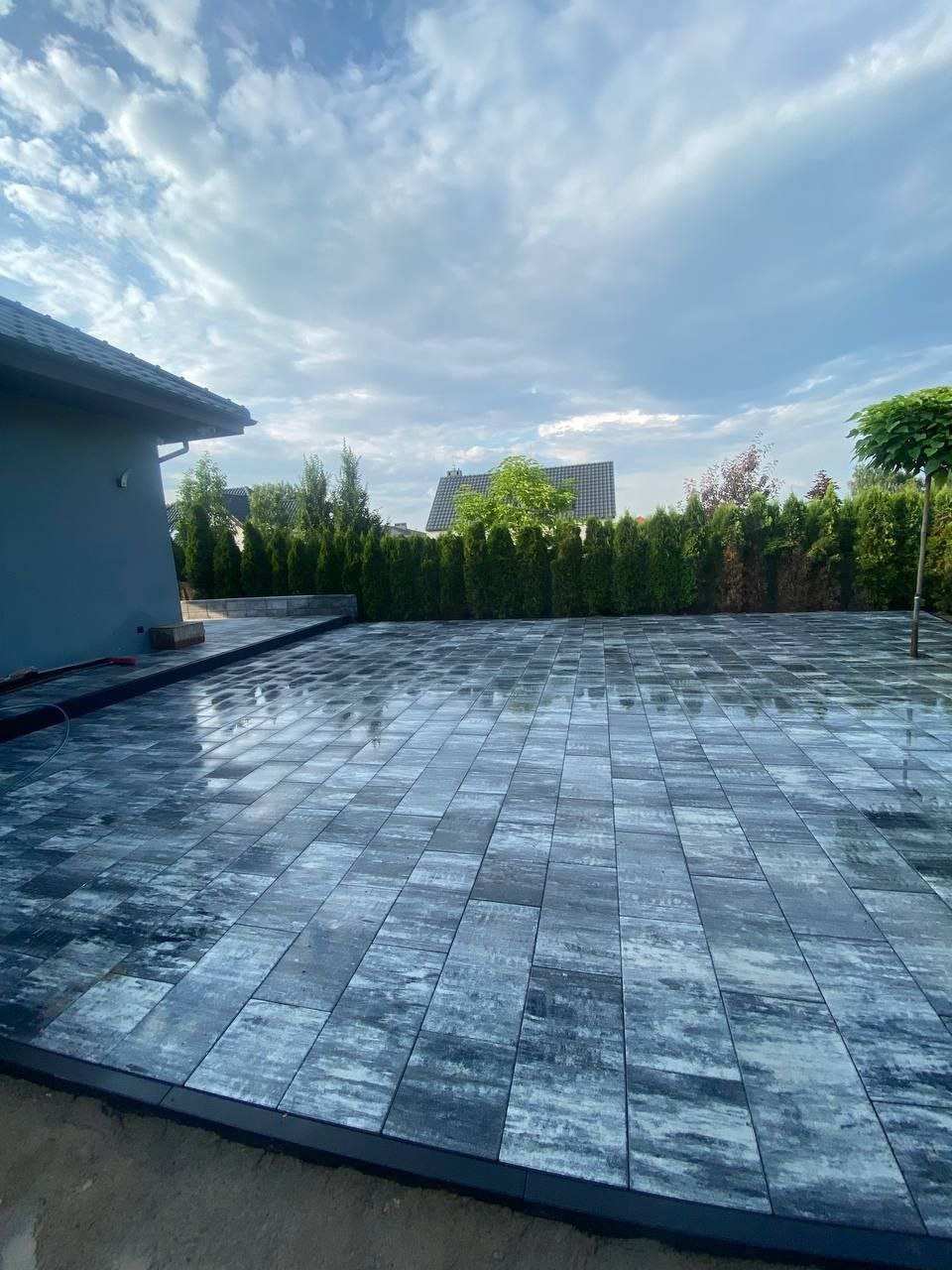 Świeżo ułożona, mokra kostka brukowa w odcieniach szarości i błękitu, odbijająca niebo. W tle zielony żywopłot i fragment domu z szarym dachem.