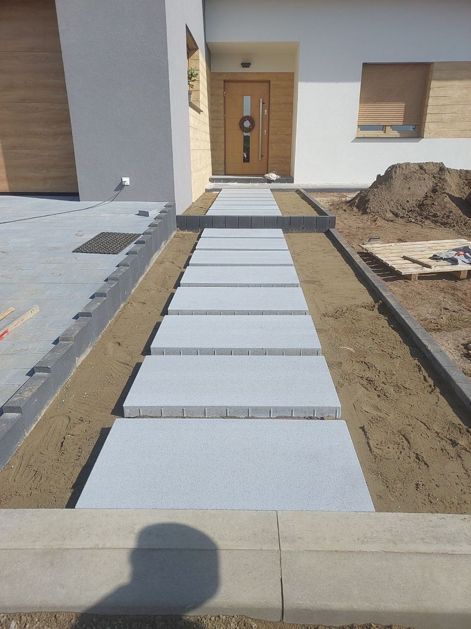 Nowoczesne podejście do domu z szarych płyt betonowych, obrzeża z kostki, widoczny fragment elewacji i drzwi wejściowe z dekoracyjnym wieńcem.