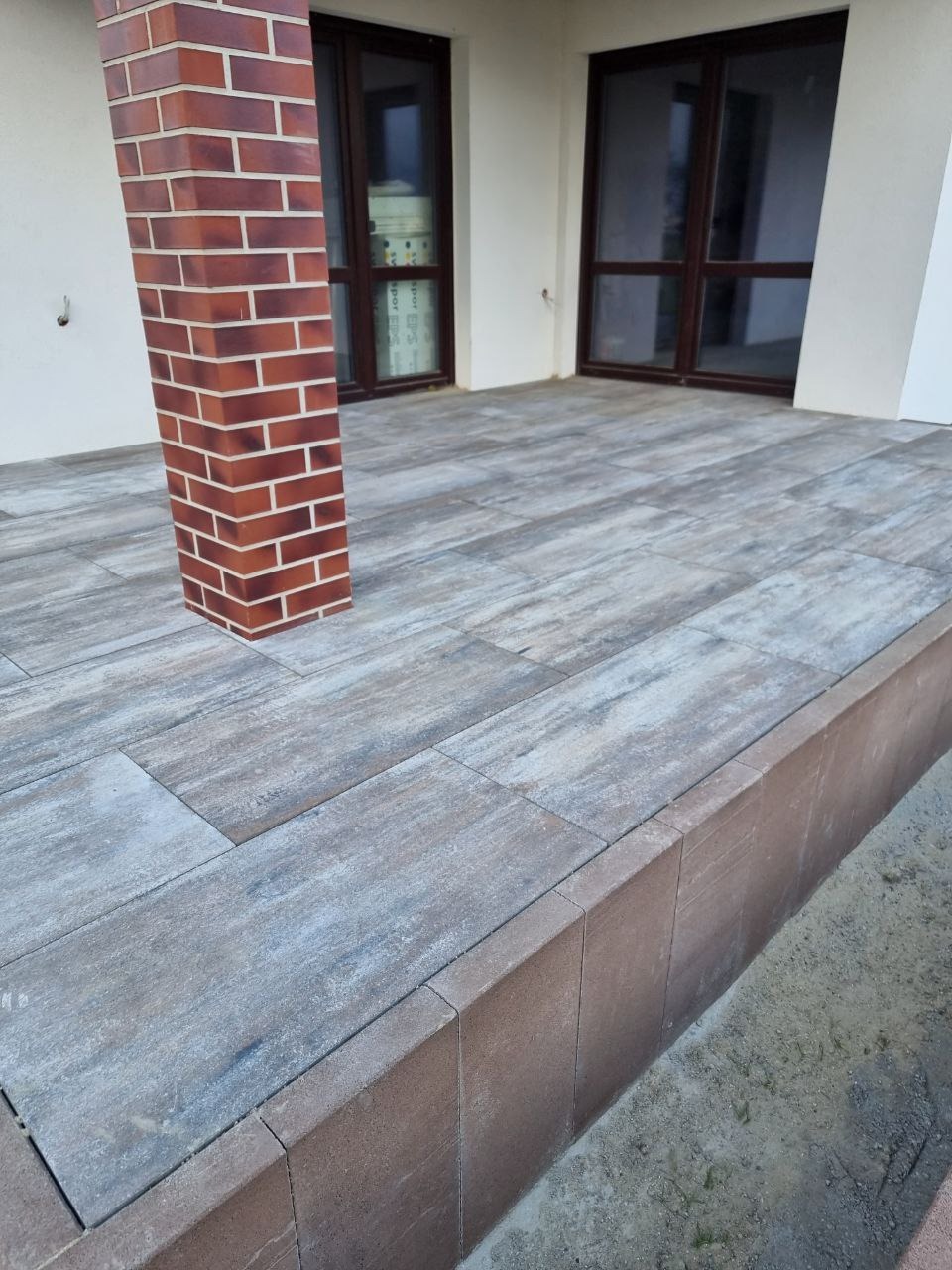 Wykończony taras z kostki brukowej w odcieniach szarości i brązu, z ceglanym filarem. Widoczne drzwi balkonowe w tle. Ujęcie z niskiej perspektywy.