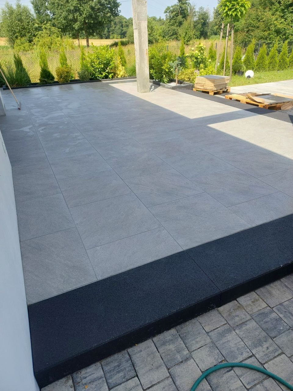 Jasnoszara, duża, kwadratowa kostka na tarasie z czarnym obrzeżem. Widoczne palety z kostką i zieleń ogrodu w tle. Ujęcie z niskiej perspektywy.