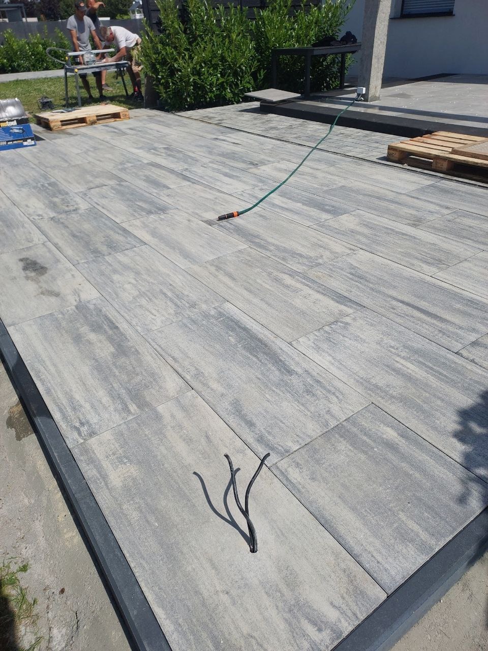 Ułożona szara kostka brukowa z widocznym wężem ogrodowym i elementami montażowymi. W tle osoby pracujące przy maszynie do cięcia kostki.