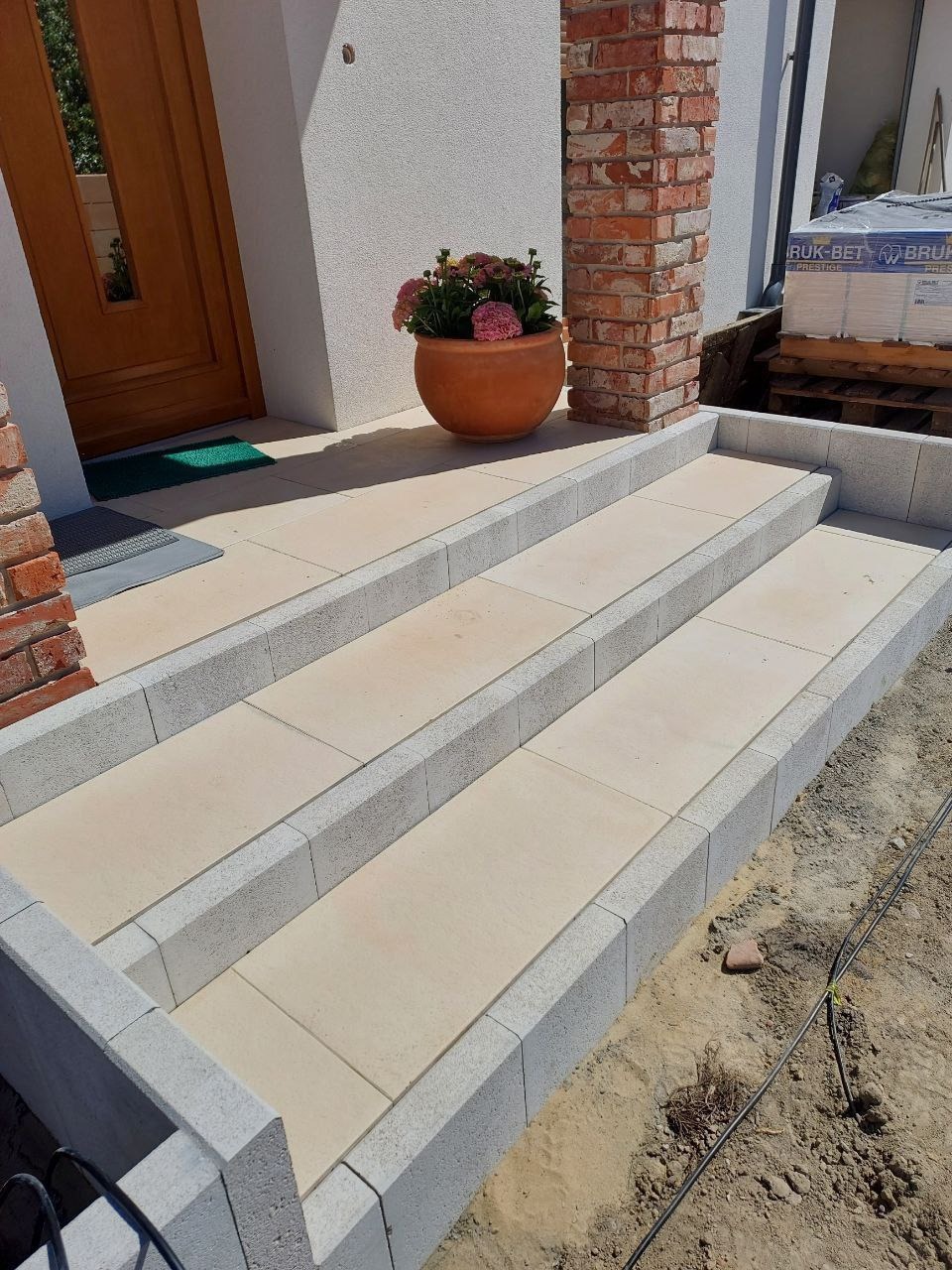 Schody wejściowe wyłożone jasną kostką brukową z szarym obramowaniem, donica z kwiatami przy drzwiach, widoczne palety z kostką w tle.