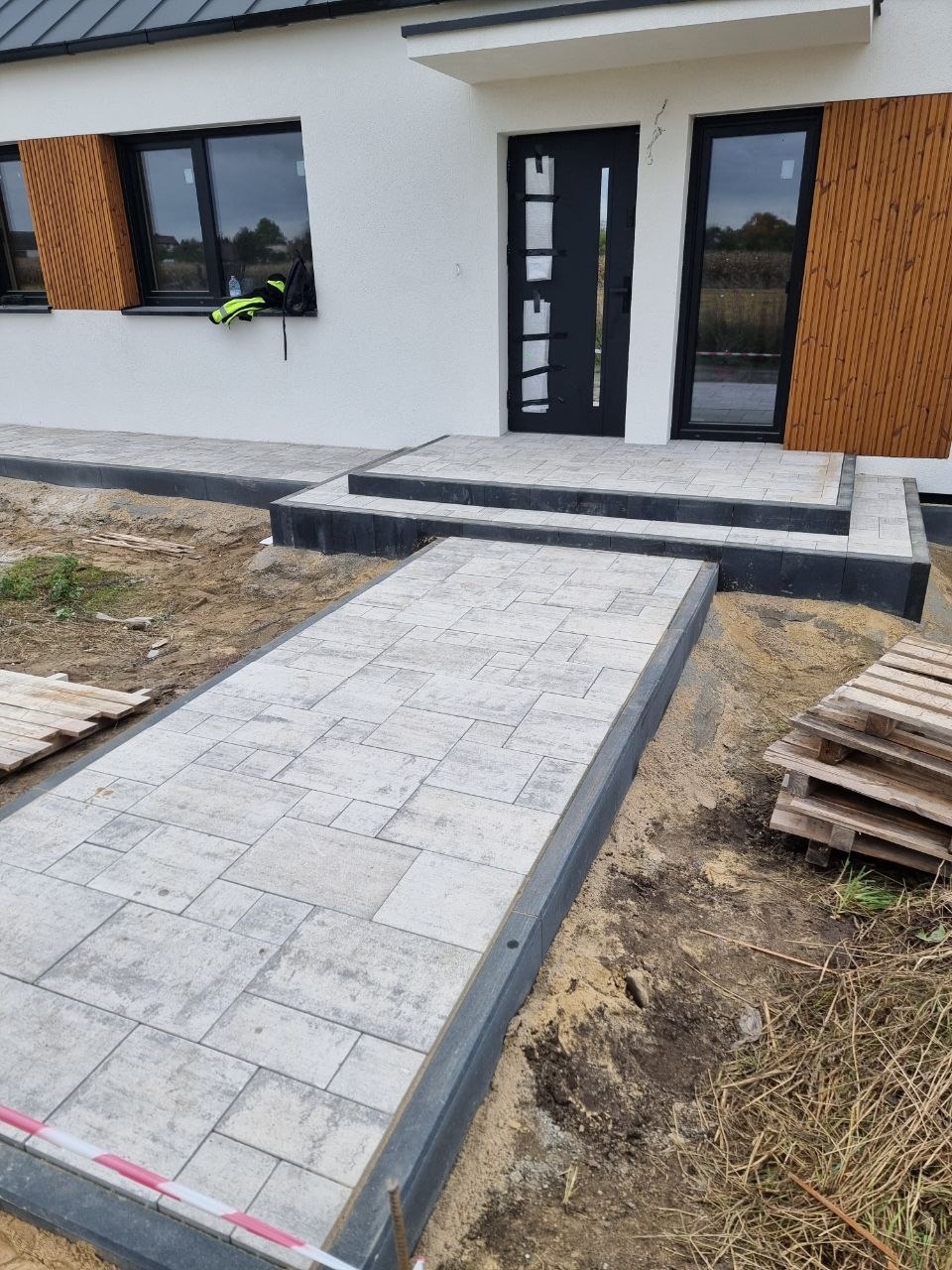 Ułożona ścieżka z kostki brukowej prowadząca do nowoczesnego domu z czarnymi oknami i drzwiami. Widoczne palety z kostką i elementy wykończeniowe.