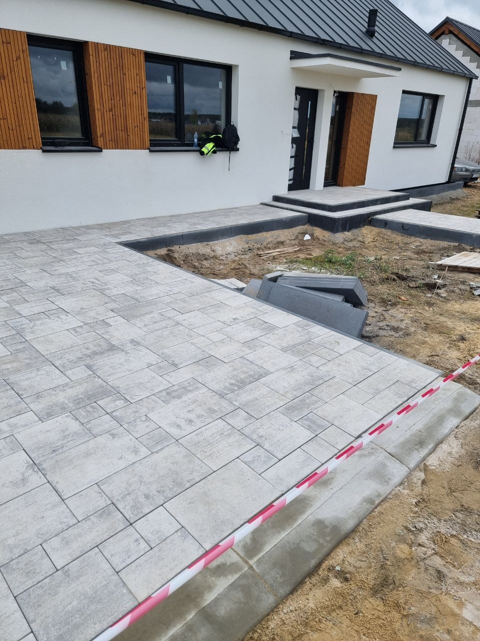 Jasnoszara kostka brukowa ułożona przed nowoczesnym domem z czarnymi oknami i drewnianymi akcentami. Częściowo ukończona praca, widoczne niedokończone krawędzie.