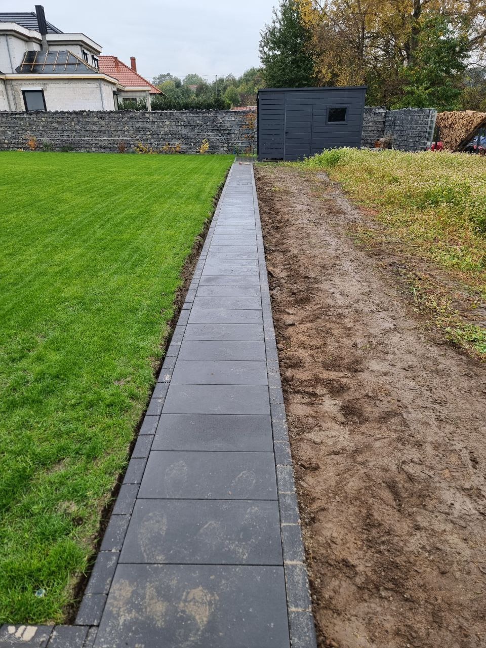 Ścieżka z ciemnej kostki brukowej oddzielająca trawnik od ziemi, z widocznym domem i szopą w tle. Ujęcie z poziomu gruntu, podkreślające linię ścieżki.
