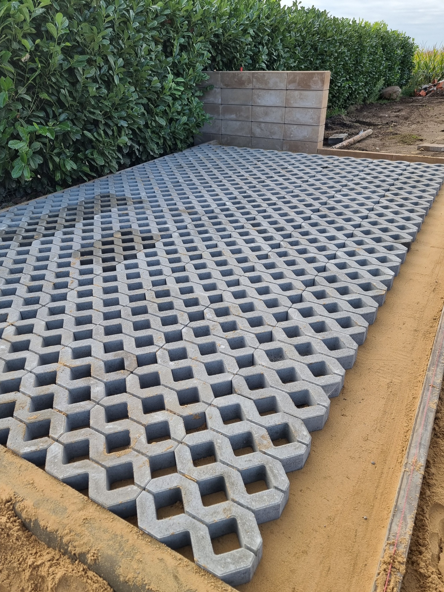 Ułożone szare ażurowe płyty betonowe na piaskowym podłożu, widoczne elementy ogrodzenia i zielony żywopłot w tle. Kątowe ujęcie z bliska.