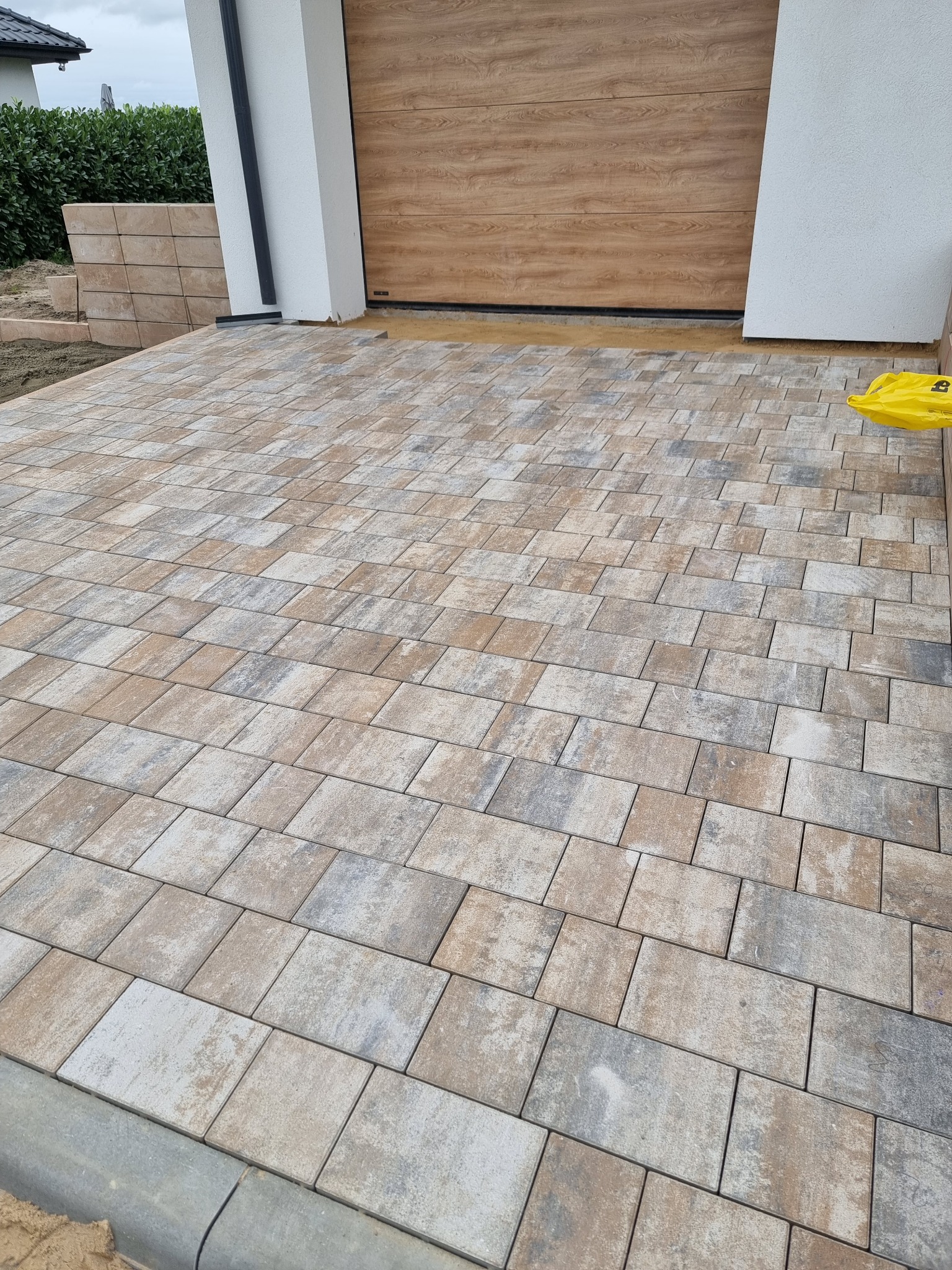 Ułożona kostka brukowa w Lesznie, prowadząca do garażu z drewnianą bramą. Widoczne nieregularne wzory i kolorystyka kostki, obrzeża oraz fragment ściany budynku.