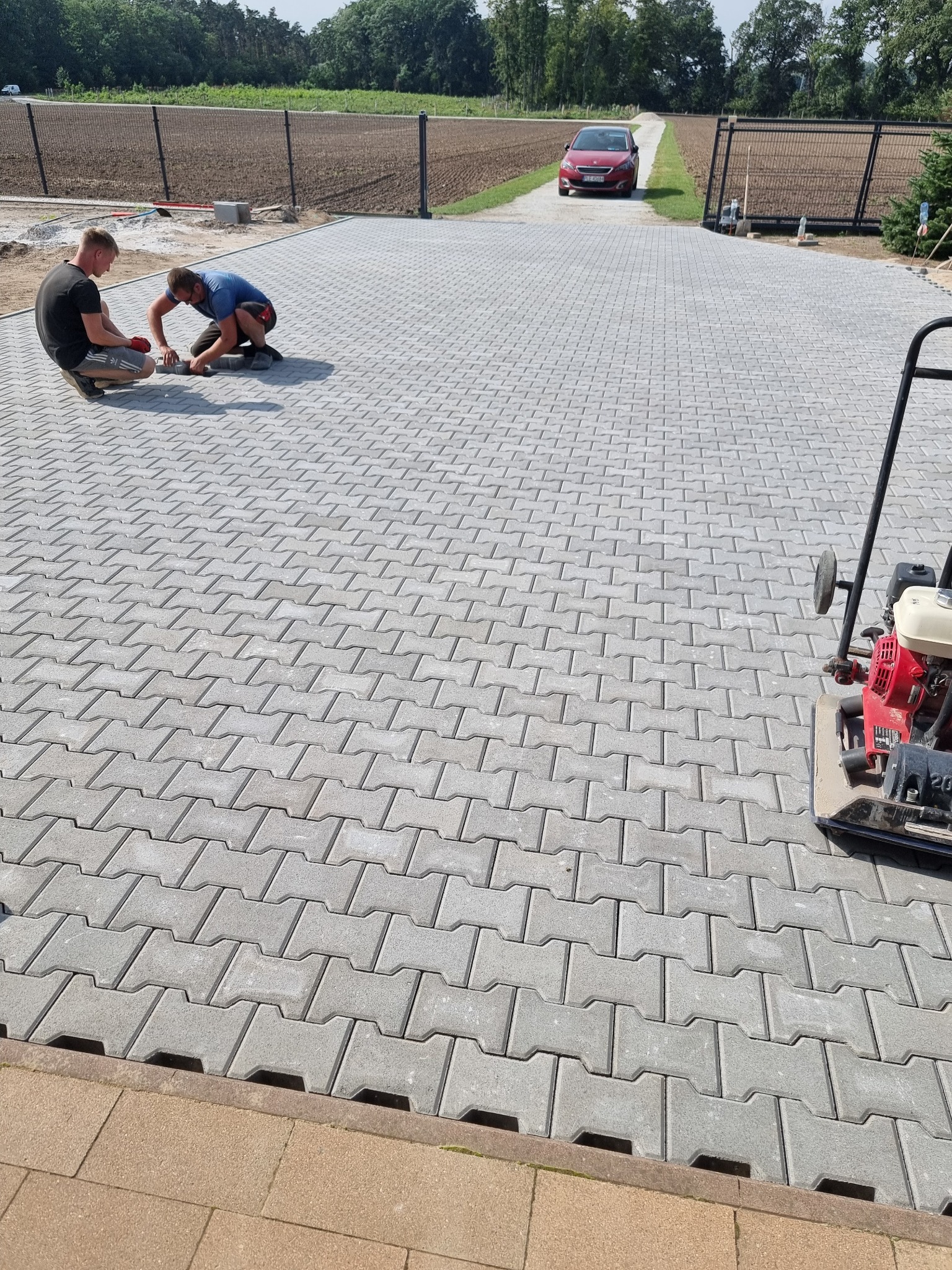 Dwóch mężczyzn układa szare kostki brukowe na podjeździe, w tle czerwony samochód i czarne ogrodzenie. Widoczna zagęszczarka.