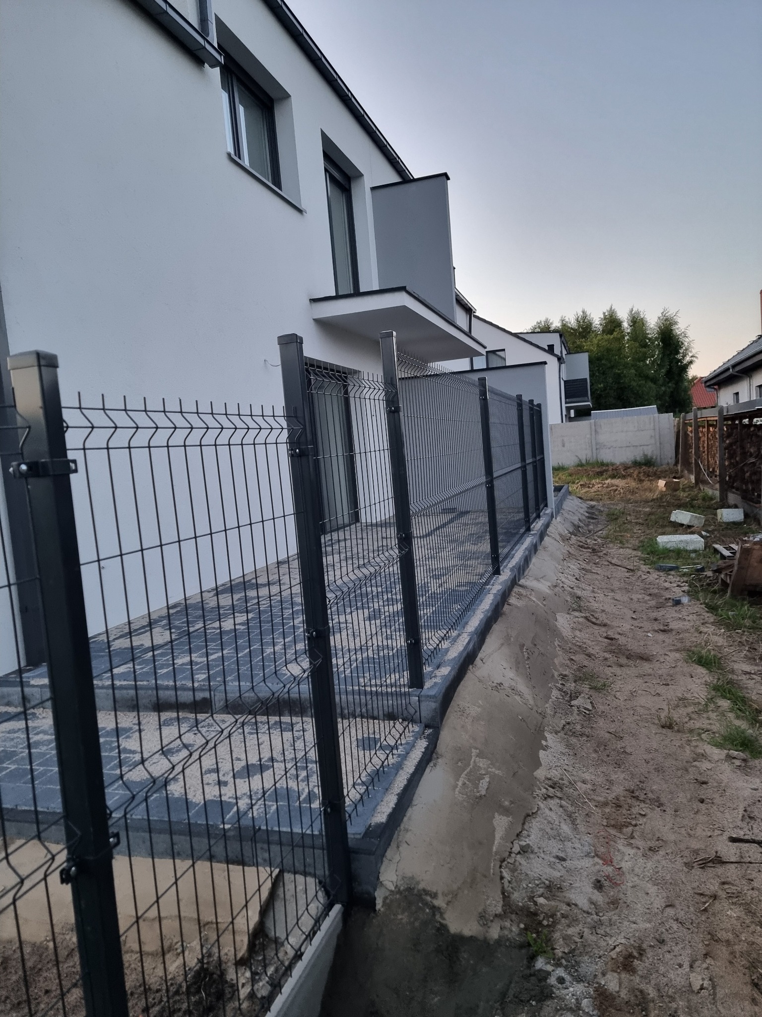 Nowoczesne ogrodzenie panelowe w kolorze grafitowym, zamontowane przy posesji z kostką brukową. Widok na dom z jasną elewacją i fragment ogrodu.