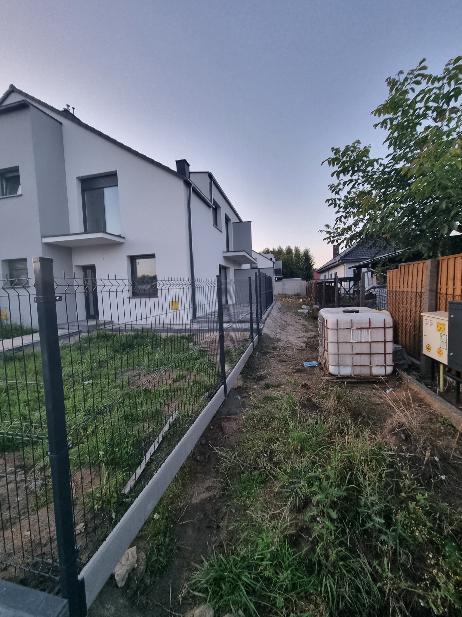 Nowoczesne ogrodzenie panelowe z podmurówką na tle domu jednorodzinnego w trakcie montażu. Widoczny zbiornik na wodę i trawa.