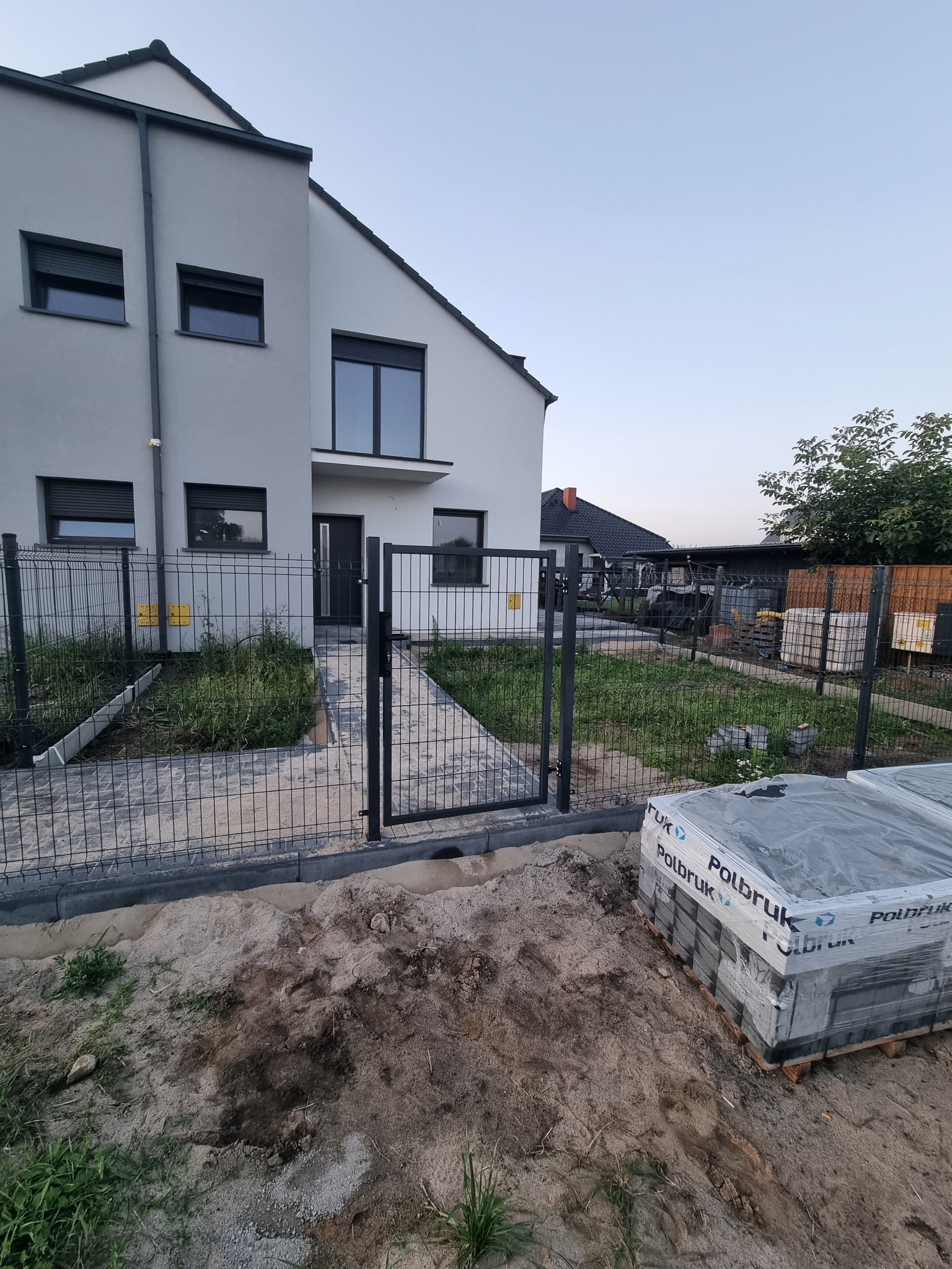 Nowoczesny dom z szarą elewacją i metalowym ogrodzeniem panelowym. Częściowo ułożona kostka brukowa i paleta kostki Polbruk. Widok na ogród i zabudowania gospodarcze.