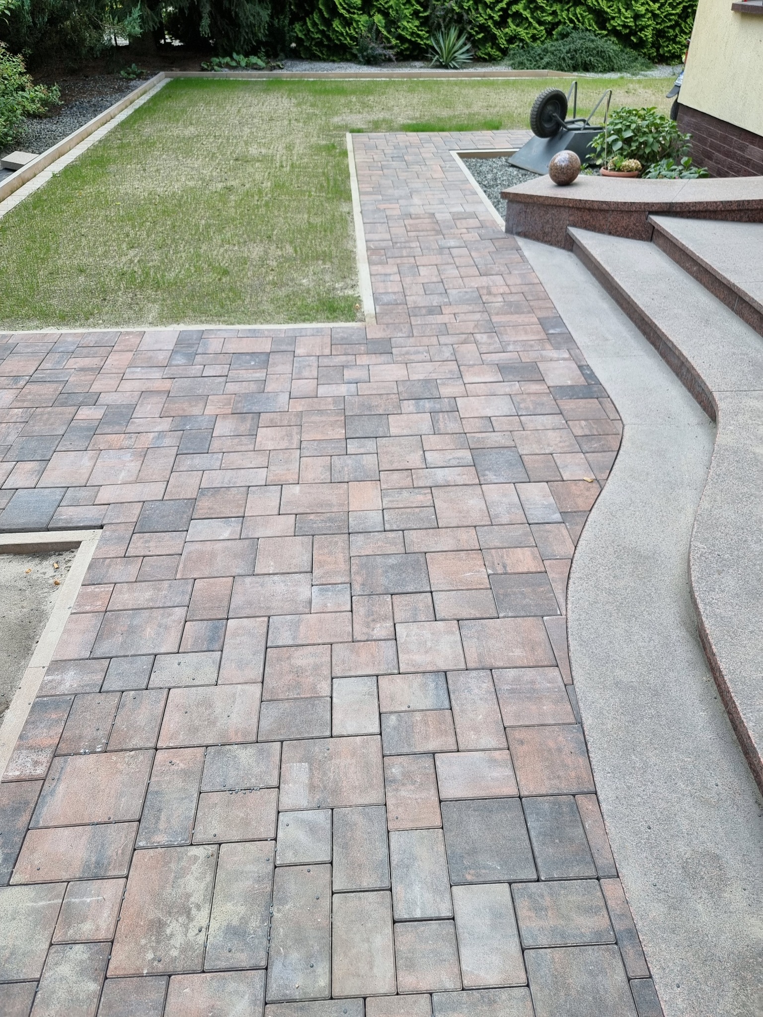 Ułożona kostka brukowa w odcieniach szarości i brązu tworzy ścieżkę w ogrodzie. Widoczne schody z szarego kamienia i trawnik z obrzeżem. W tle zieleń drzew i krzewów.