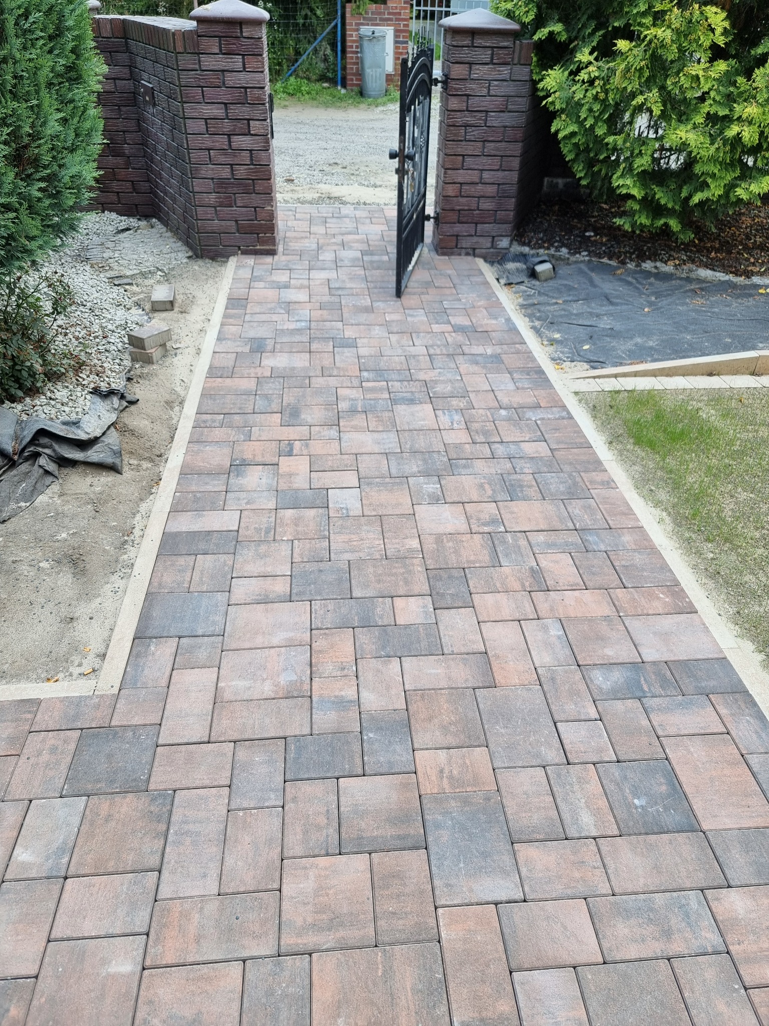Ścieżka z kostki brukowej w odcieniach brązu i szarości, prowadząca przez bramę z ceglanymi słupkami. Widoczne elementy ogrodu i przygotowane podłoże pod dalsze prace.