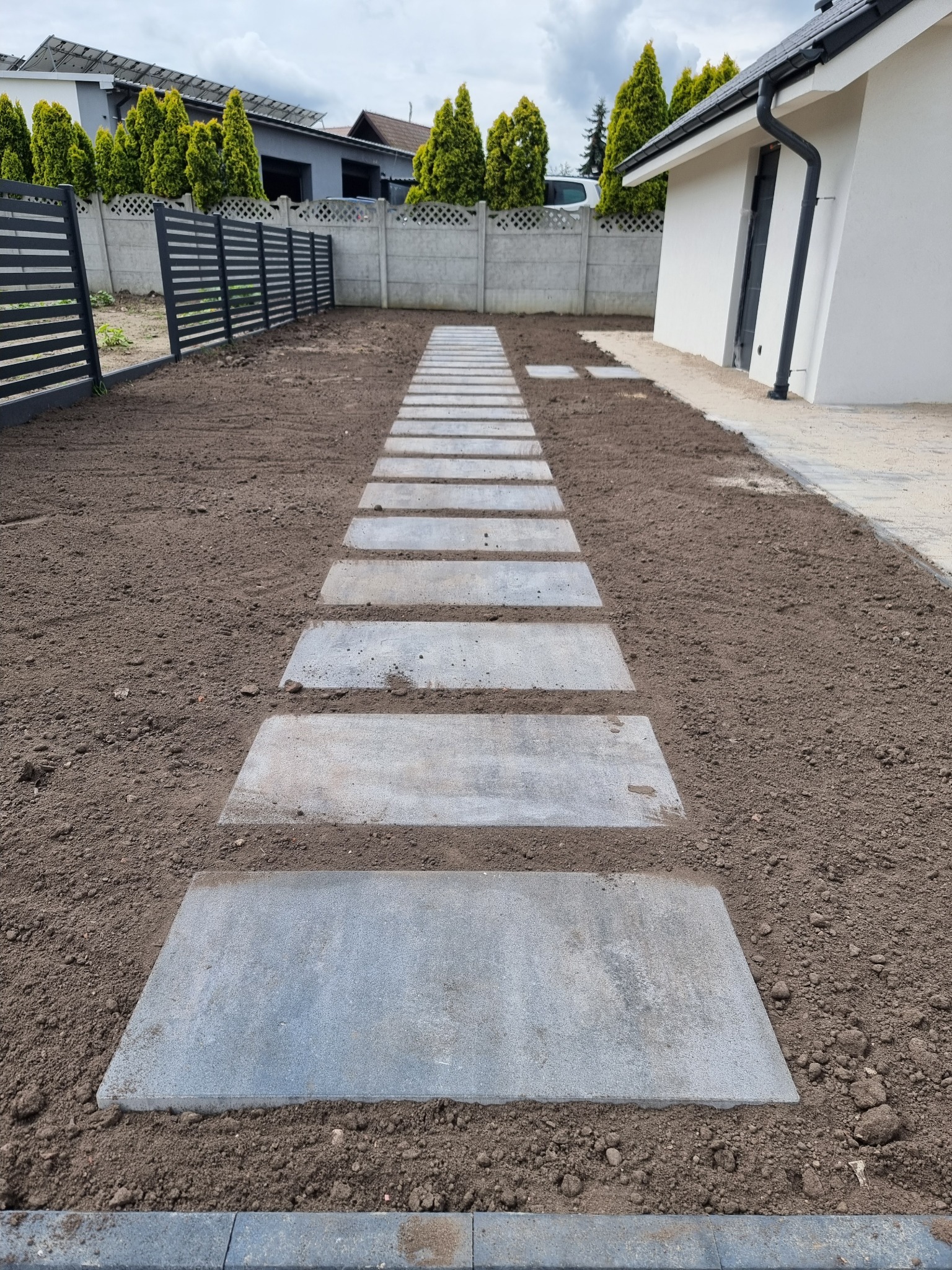 Ścieżka z płyt betonowych w ogrodzie, świeżo obsypana ziemią, z widocznym ogrodzeniem panelowym i fragmentem budynku w tle. Ujęcie z perspektywy.