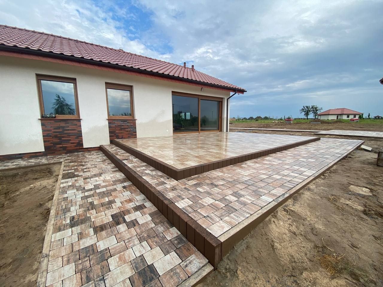 Nowoczesny taras i ścieżka z kostki brukowej w odcieniach brązu, przylegające do domu z jasną elewacją i brązowymi akcentami. Widok na ogród i pochmurne niebo.