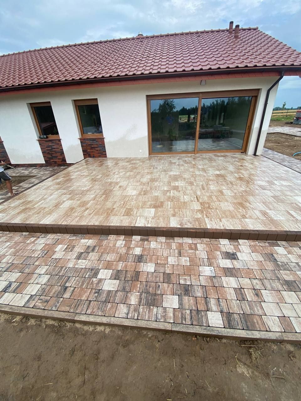 Wykończony taras z kostki brukowej w odcieniach brązu i beżu, przylegający do domu z czerwonym dachem. Widoczne schody z kostki i fragment trawnika.