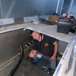 Aleks Tuszyński Usługi Elektryczne - Elektryk w trakcie pracy w pomieszczeniu technicznym, widoczne rury i narzędzia. Uwagę zwraca specyficzne oświetlenie i metalowe wykończenie ścian.