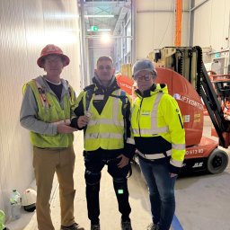 Aleks Tuszyński Usługi Elektryczne - Trzech elektryków w odblaskowych ubraniach pozuje na tle podnośnika i instalacji kablowych w hali. Jeden z nich trzyma odznakę.