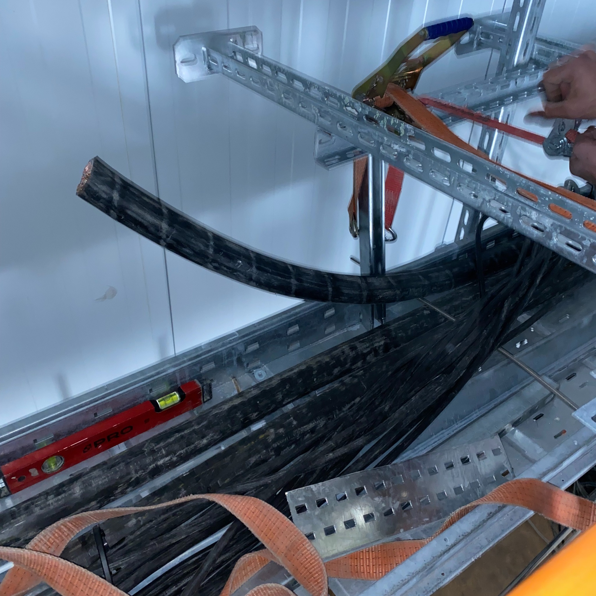 Kable elektryczne ułożone w metalowych korytach, zabezpieczone pasami transportowymi. Widoczna poziomica i narzędzia monterskie.