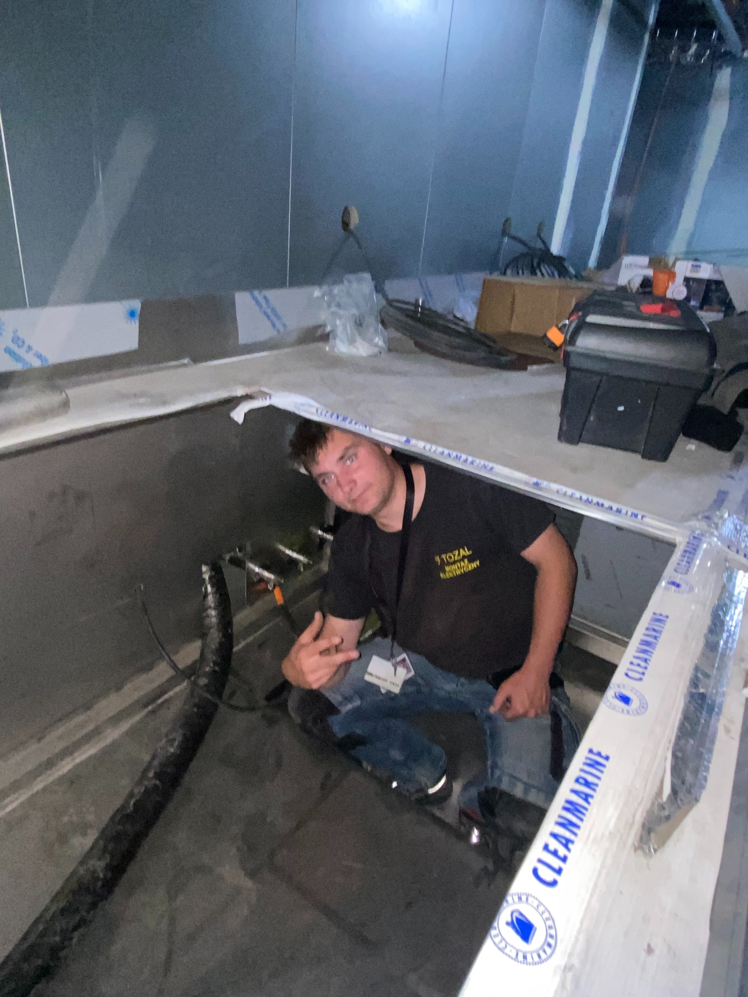 Elektryk w trakcie pracy w pomieszczeniu technicznym, widoczne rury i narzędzia. Uwagę zwraca specyficzne oświetlenie i metalowe wykończenie ścian.