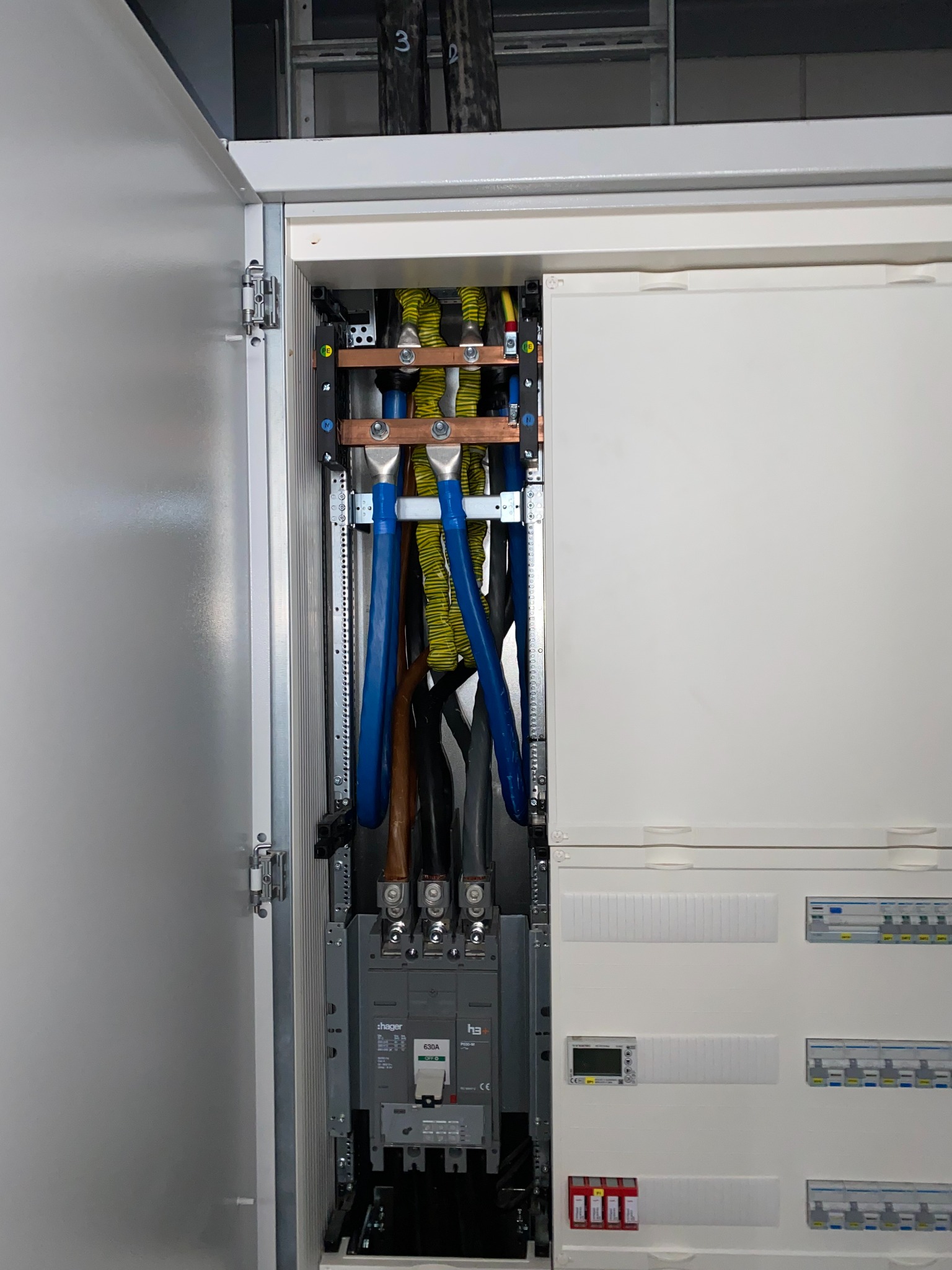 Otwarta szafa elektryczna z widocznymi przewodami w izolacji żółto-czarnej i niebieskiej, zabezpieczeniami Hager i elementami montażowymi. Widok z bliska na wnętrze rozdzielnicy.