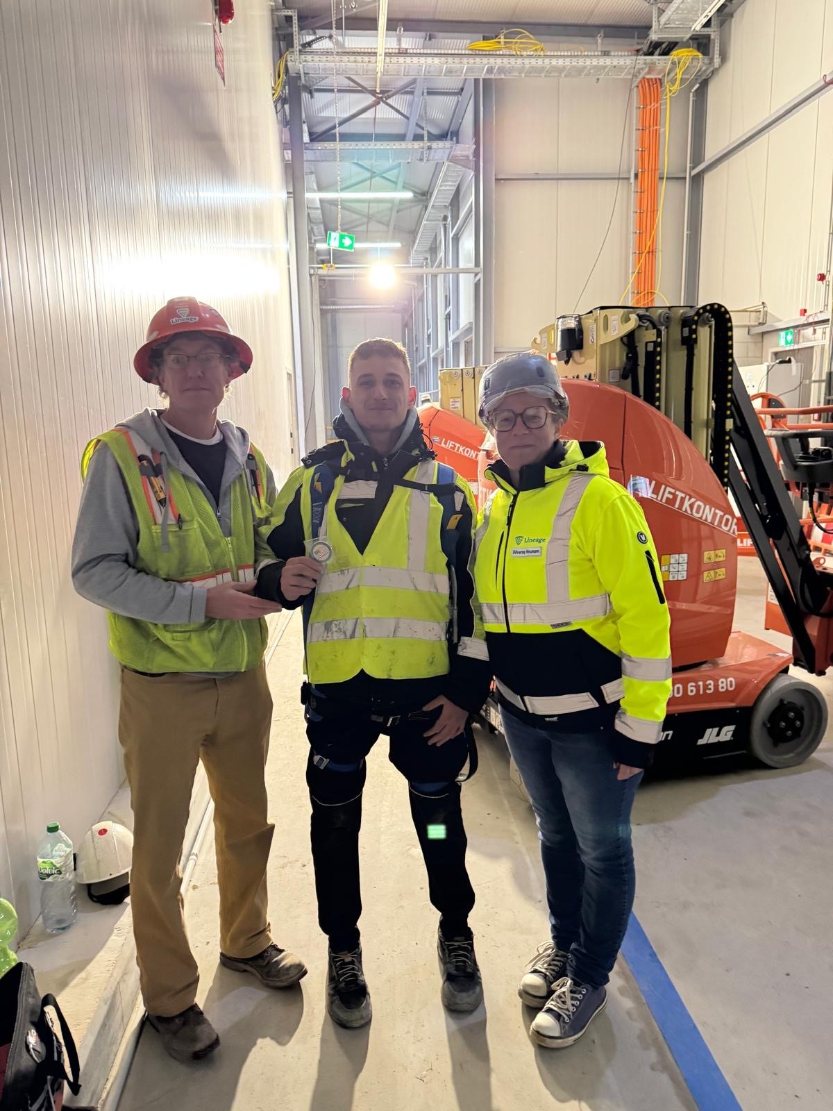 Trzech elektryków w odblaskowych ubraniach pozuje na tle podnośnika i instalacji kablowych w hali. Jeden z nich trzyma odznakę.