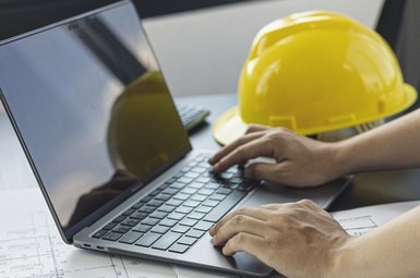 Praca przy laptopie z żółtym kaskiem w tle. Ręce na klawiaturze, widoczne plany architektoniczne. Kadr z bliska, oświetlenie biurowe.