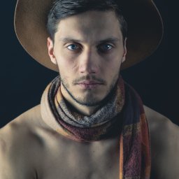 Marcin Pycha Studio - Portret mężczyzny w kapeluszu i szaliku na nagim ciele, z intensywnym spojrzeniem i ciemnym tłem, ukazujący artystyczny styl i dbałość o detale.