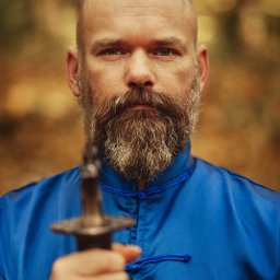 Marcin Pycha Studio - Portret mężczyzny z brodą w niebieskim stroju, trzymającego miecz na tle jesiennego lasu. Ujęcie z bliska, ostre światło podkreśla detale twarzy.