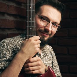 Marcin Pycha Studio - Uśmiechnięty mężczyzna w okularach, opierający się o czerwoną gitarę na tle ceglanej ściany. Portret muzyka z instrumentem w studiu.