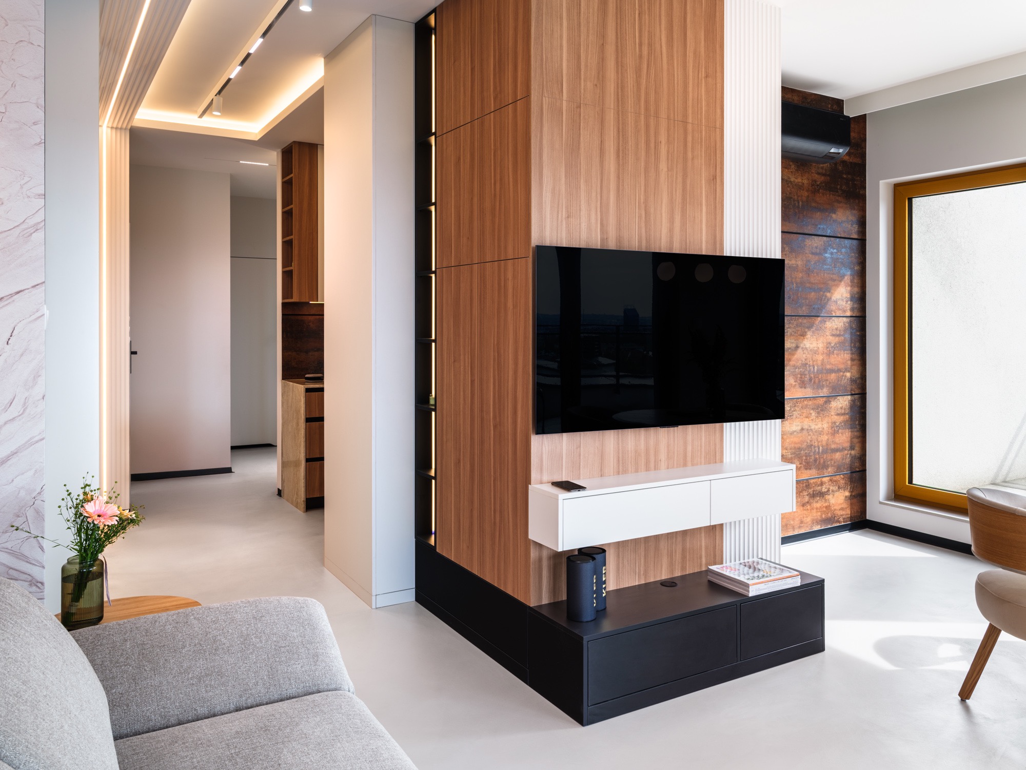 Nowoczesne wnętrze: drewniana ściana z TV, białe elementy, szara sofa i kwiaty. Minimalistyczny design z oświetleniem LED i otwartą przestrzenią.