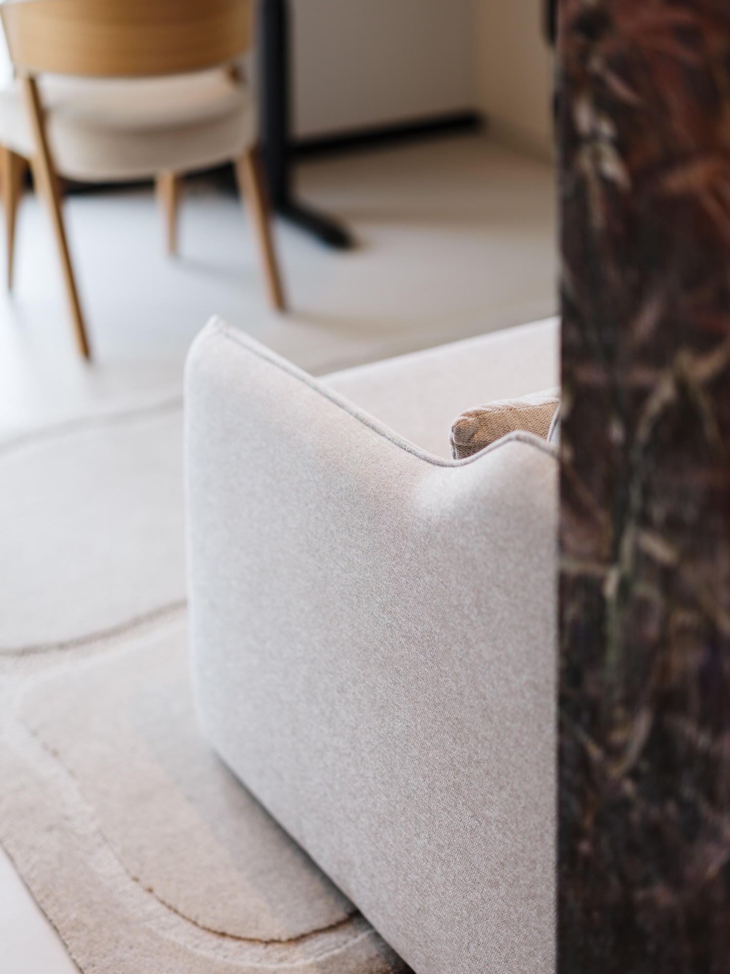 Jasny, minimalistyczny salon z beżową sofą i dywanem w tonacji. W tle krzesło i stół. Część kadru zasłania dekoracyjny panel ścienny.