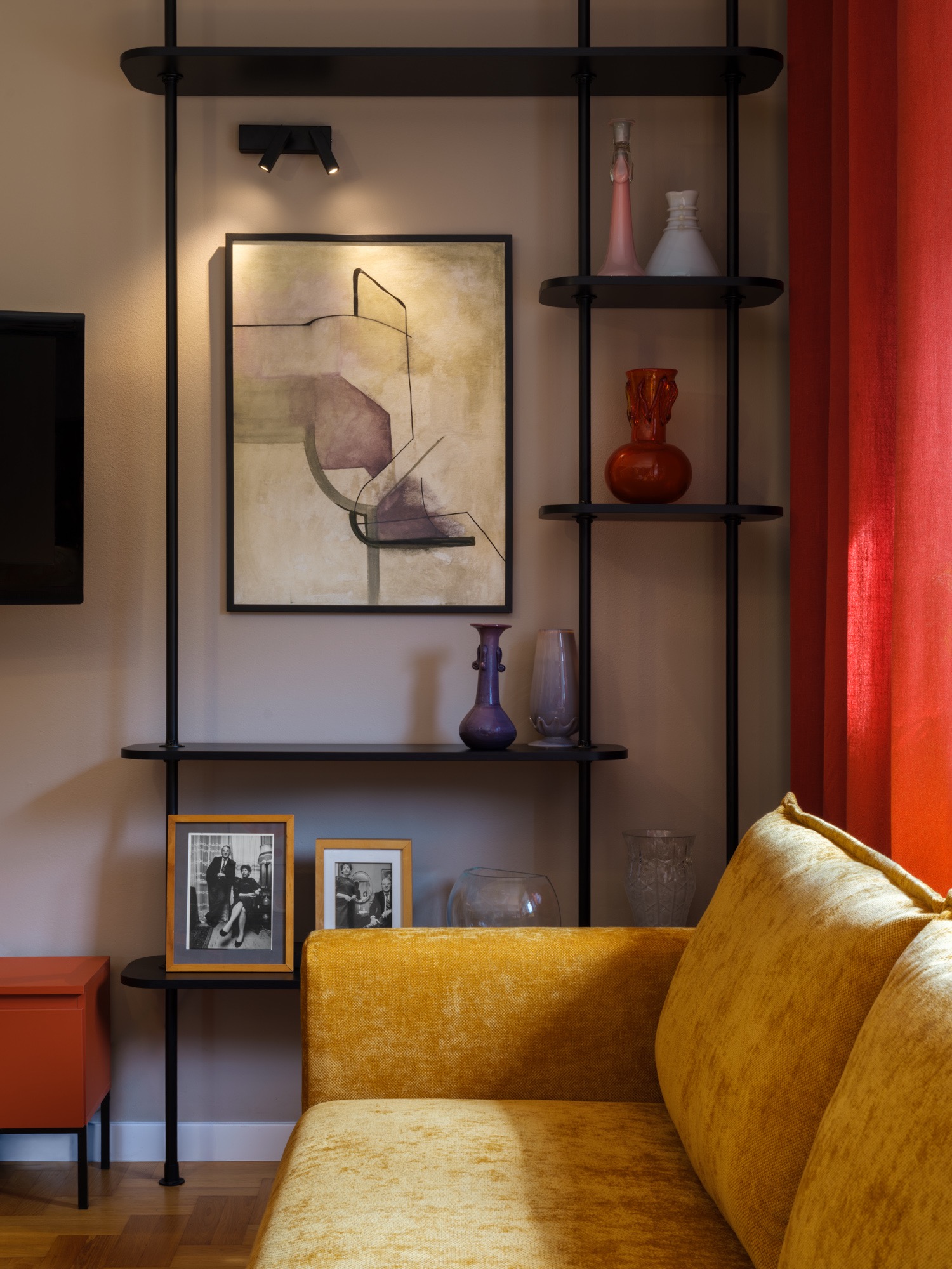 Stylowe wnętrze: żółta sofa z poduszkami, czarny regał z dekoracjami (wazony, zdjęcia w ramkach) i abstrakcyjny obraz na ścianie. Czerwona zasłona dodaje koloru.