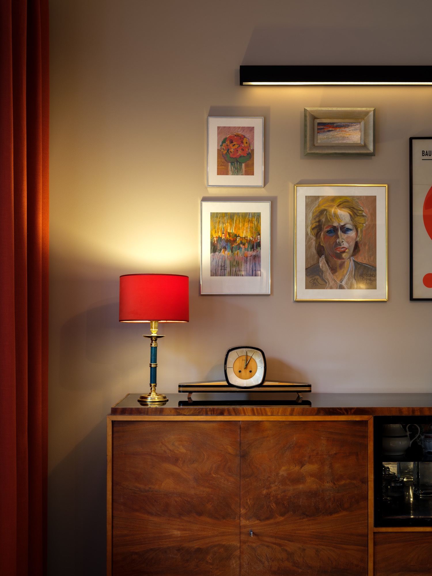 Stylowe wnętrze: komoda z forniru, lampa z czerwonym abażurem, zegar i galeria obrazów na ścianie w ciepłych barwach. Akcent stanowi pomarańczowa zasłona.