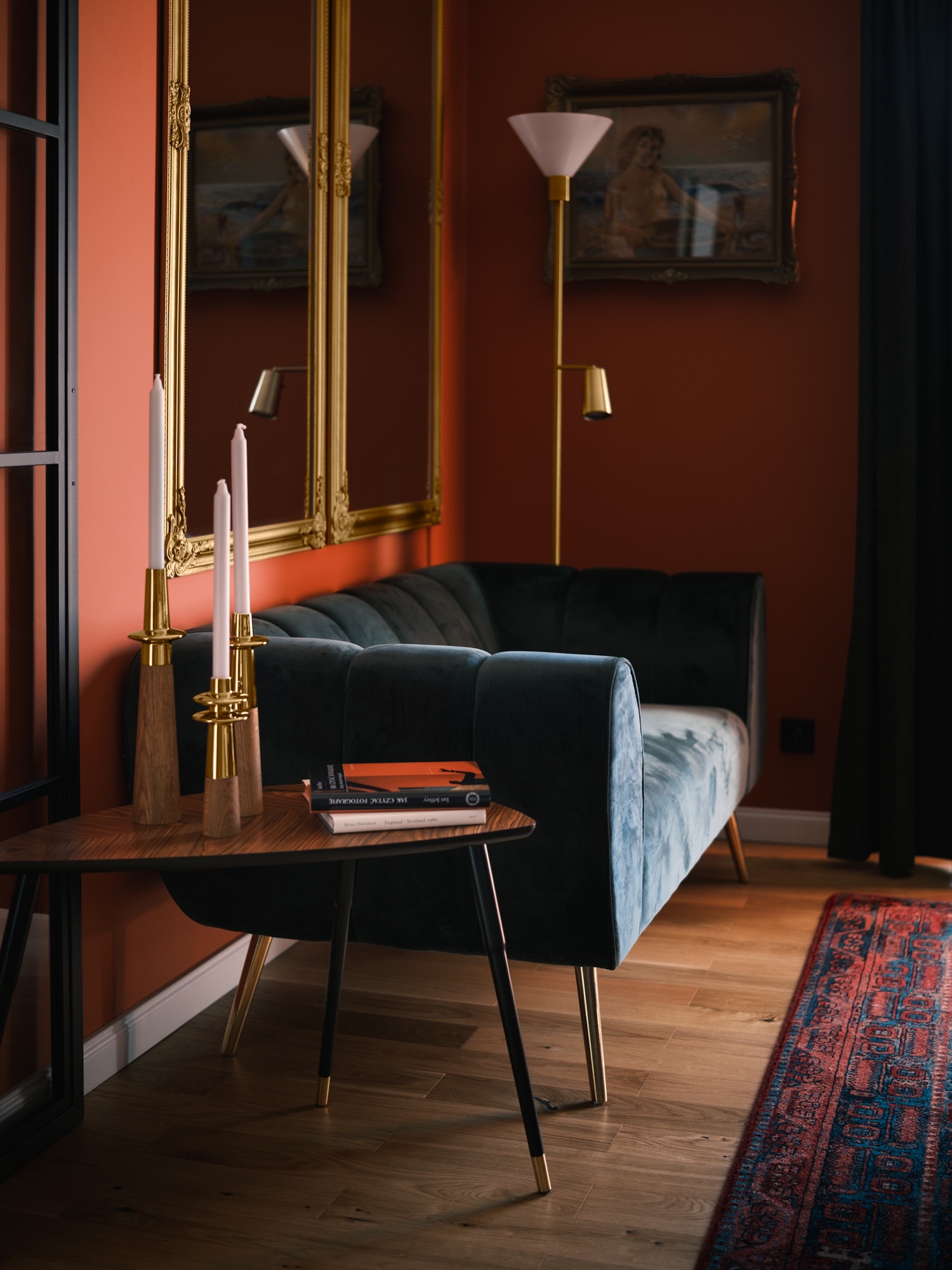 Stylowe wnętrze z turkusową sofą, stolikiem z książkami i świecznikami. W tle lustro w złotej ramie i obraz na ścianie w odcieniach terakoty. Parkiet i dywan.