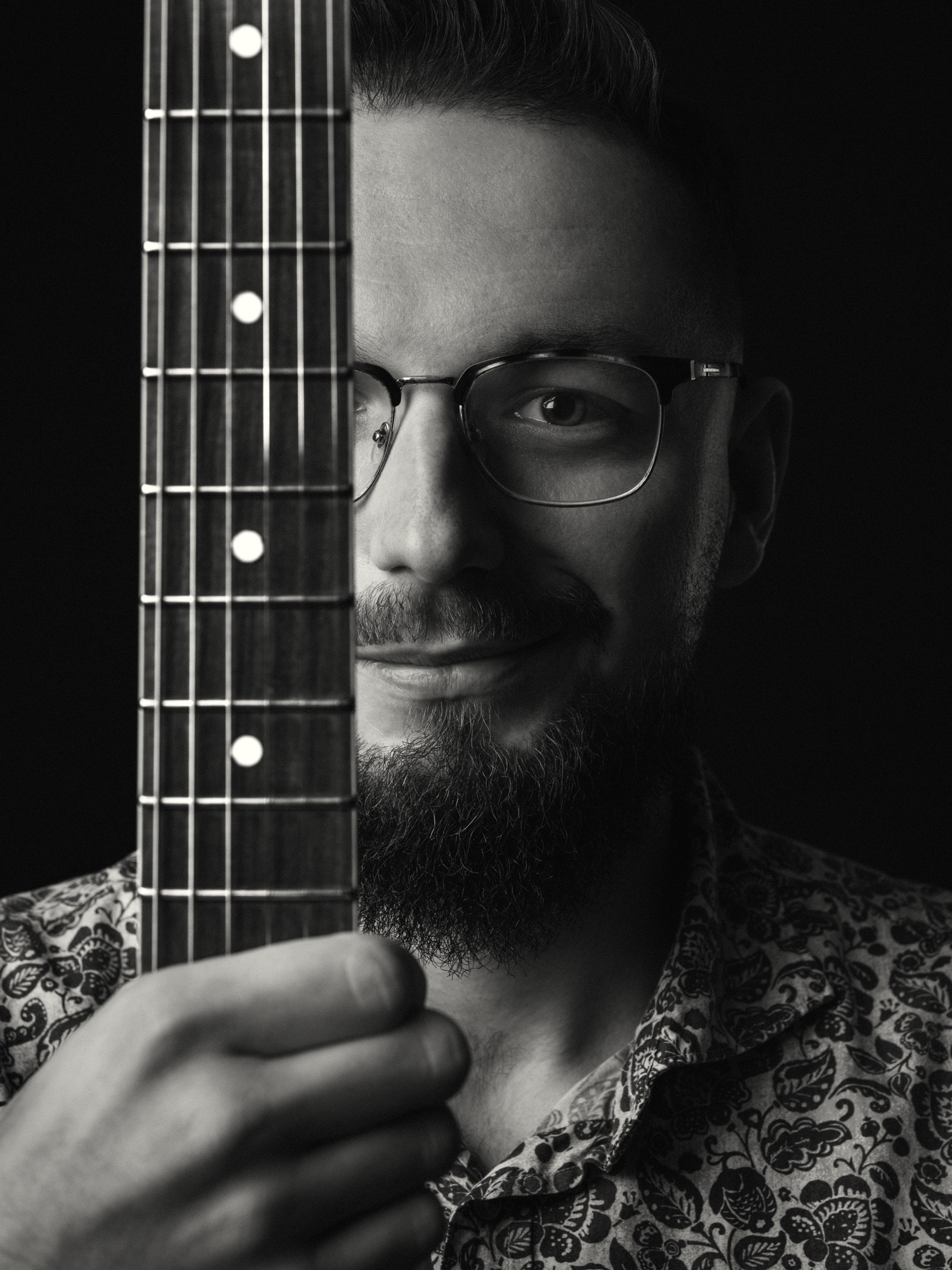Czarno-biały portret brodatego mężczyzny w okularach, z gryfem gitary zasłaniającym część twarzy, na ciemnym tle. Ubrany w koszulę we wzory.