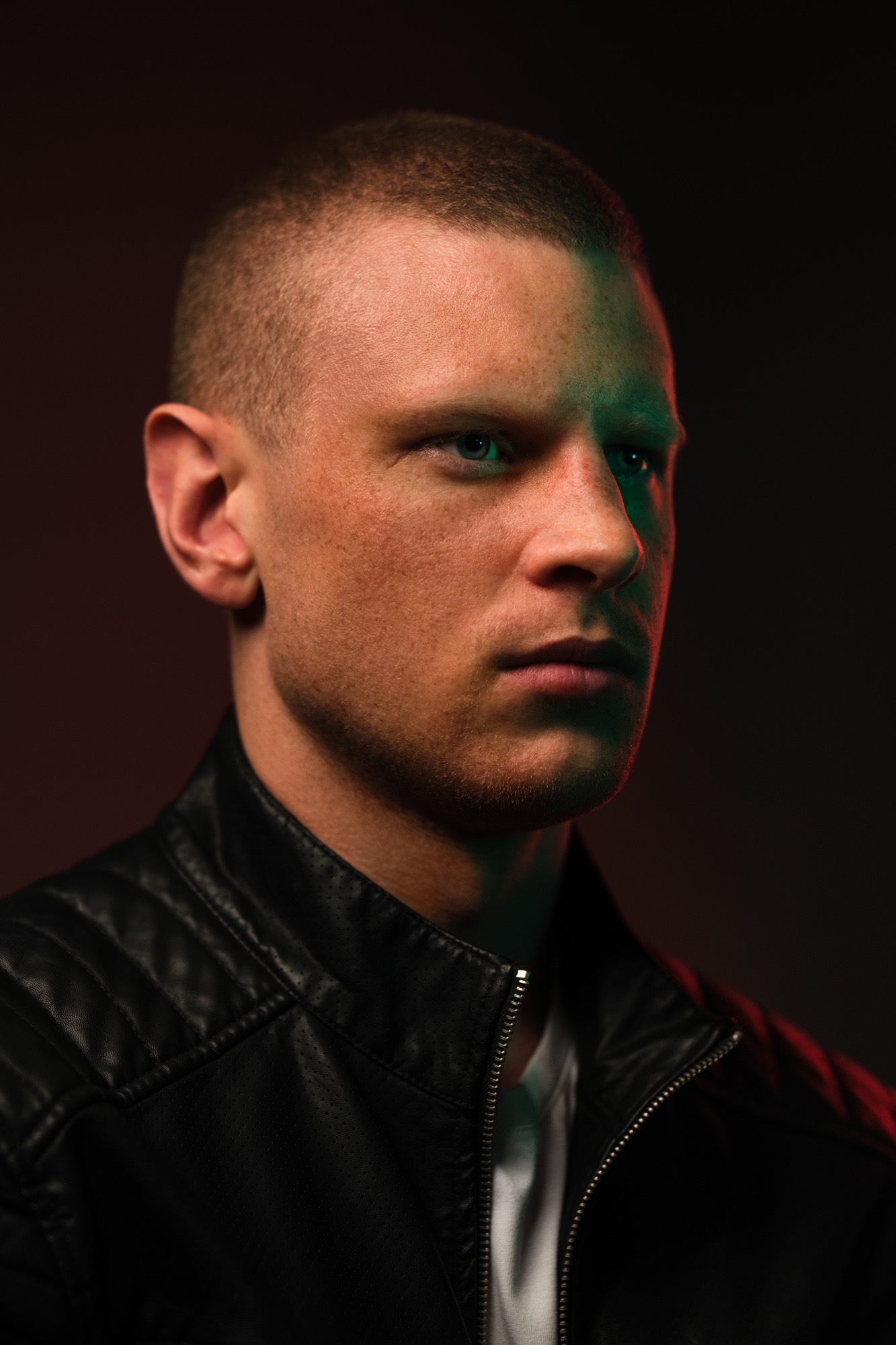 Portret mężczyzny w czarnej skórzanej kurtce, oświetlonego zielonym i czerwonym światłem, z krótkimi włosami i poważnym wyrazem twarzy, na ciemnym tle.