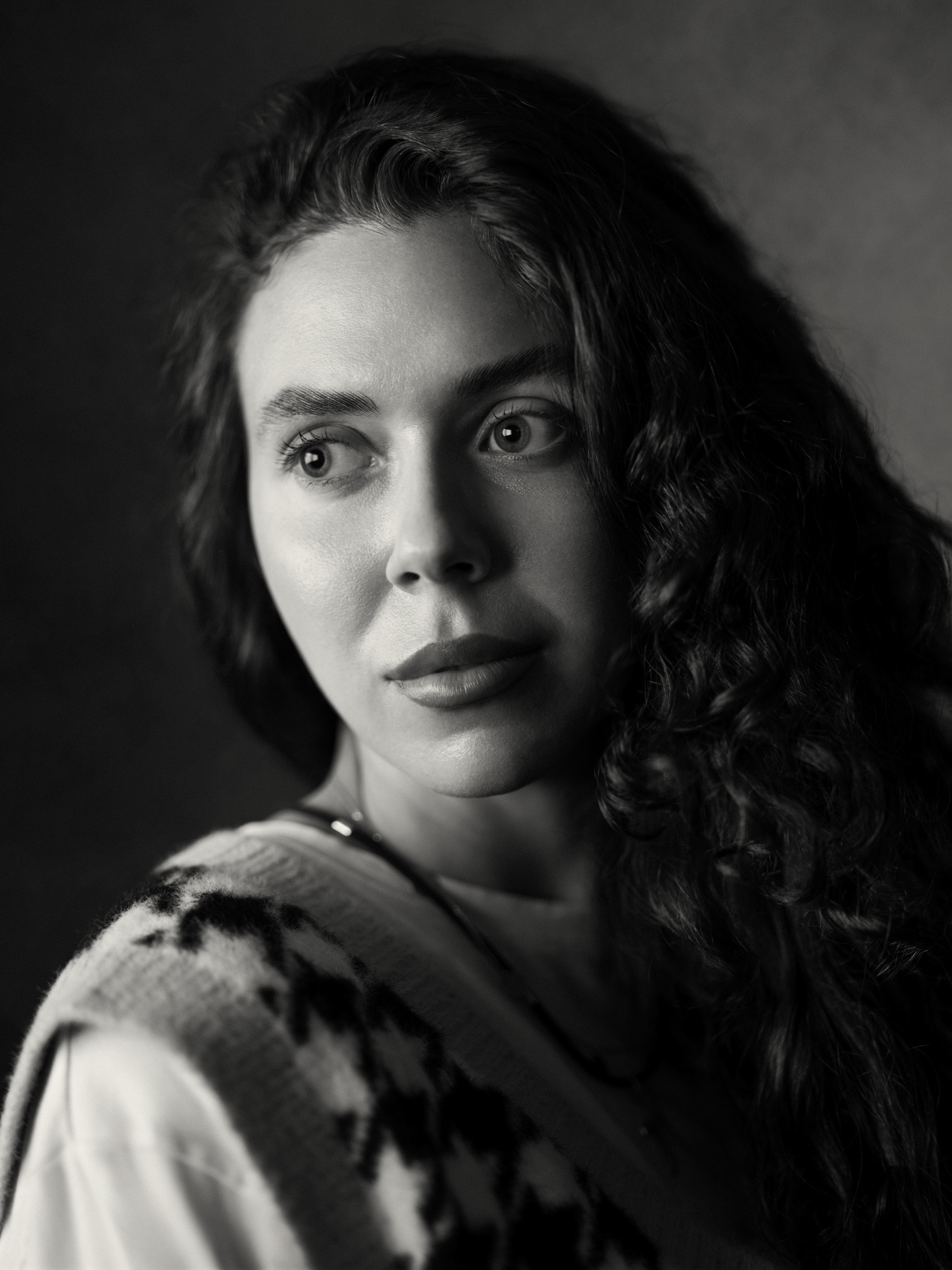 Czarno-biały portret kobiety z kręconymi włosami, spoglądającej w lewo, z delikatnym uśmiechem i wzorem na swetrze. Subtelne oświetlenie podkreśla rysy twarzy.